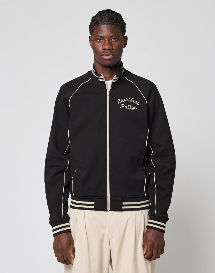 A man stands facing forward, wearing the Live Fast Tracksuit Jacket—a black zip-up with white piping, Eingrifftaschen, and a Piste Face Rallye Chainstitch Logo on the chest—paired with light pants against a plain light background.