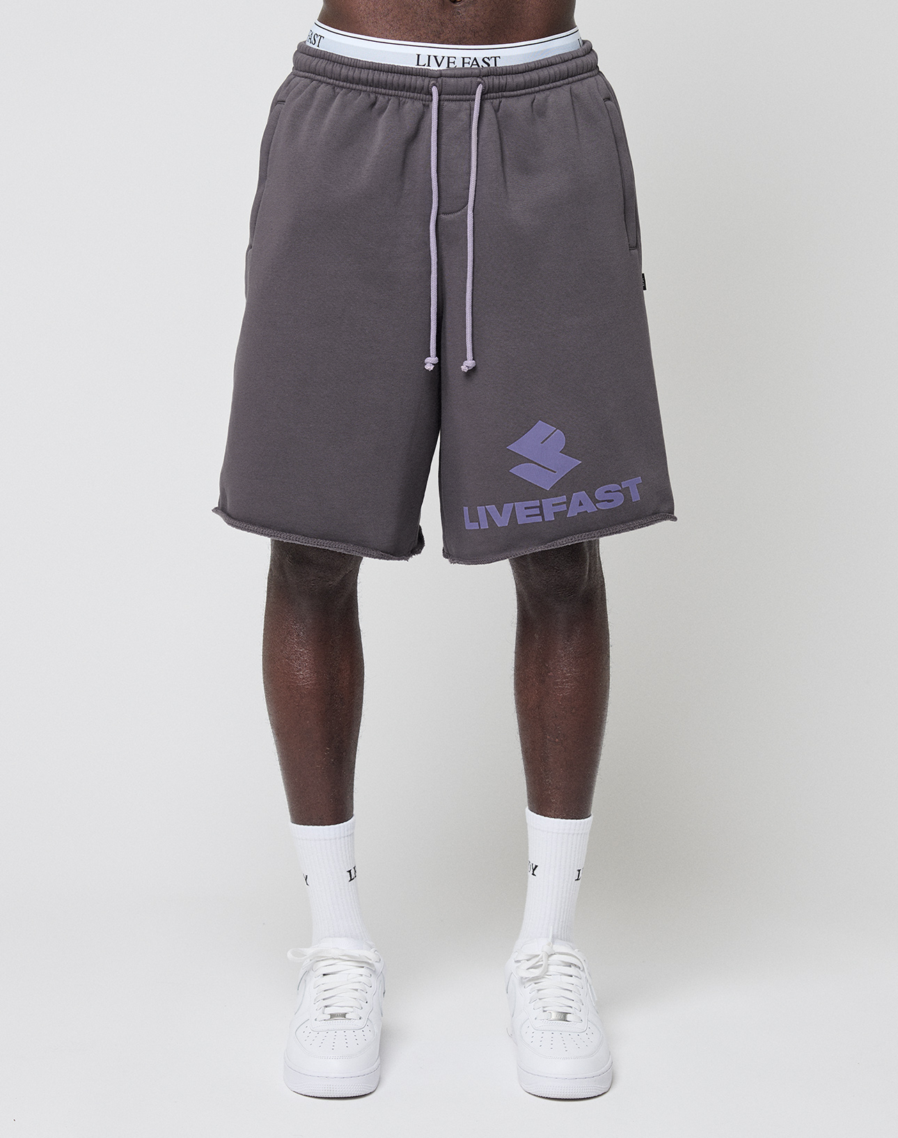 A person wears the LIVE FAST DIE YOUNG Live Fast LF Shorts in dark gray with a purple LIVEFAST logo, elastic waistband, and side pockets, paired with white socks and sneakers against a plain white background.