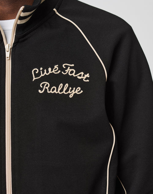A close-up of the Live Fast Tracksuit Jacket in black with beige piping, Eingrifftaschen, and Live Fast Rallye chainstitched in cursive on the left chest.
