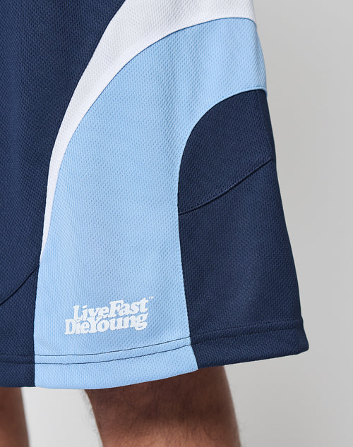 Close-up of a person wearing LIVE FAST DIE YOUNG Basic Trackshorts in navy blue with light blue and white curved panels, logo print on the lower left, and an elastic waistband for comfort.