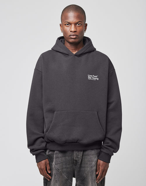 A person faces forward in an oversize fit black Live Fast NY Hooded by LIVE FAST DIE YOUNG (70% Baumwolle), featuring small white Live Fast Die Young text on the left chest, dark jeans, and a plain light background.