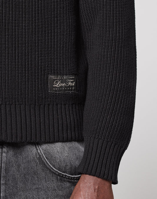 A close-up shows the lower torso and arm of a person wearing the LFDY Knit Jacket by LIVE FAST DIE YOUNG, featuring ribbed fabric, raglan sleeves, and a small "Live Fast" label near the hem, paired with faded gray jeans.
