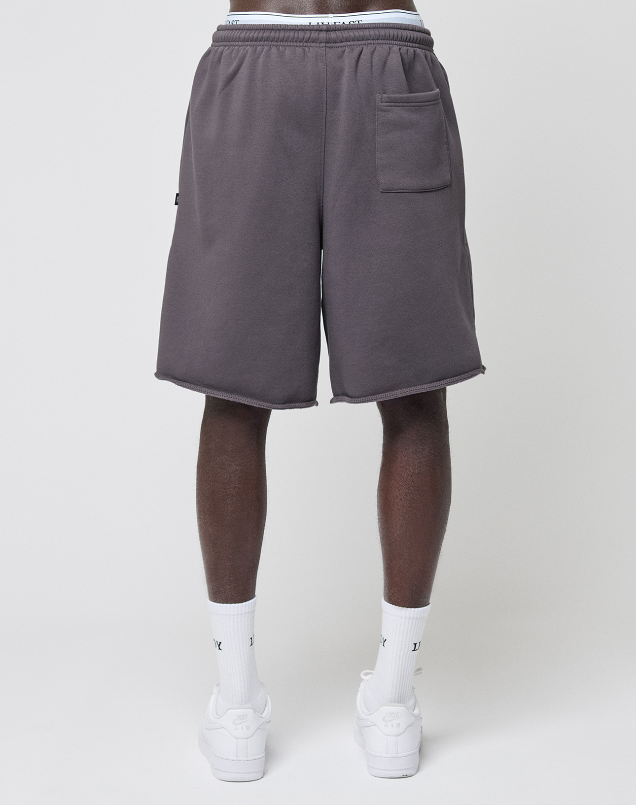 A person faces away from the camera wearing wide fit, dark gray Live Fast LF Shorts by LIVE FAST DIE YOUNG with elastic waistband and side pockets, paired with white socks and sneakers against a plain white background.