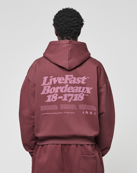 A person in an oversized maroon “Basic 360 Hooded” by LIVE FAST DIE YOUNG, featuring a bold “LiveFast Bordeaux 18-1718” backprint, stands against a plain light background facing away from the camera.