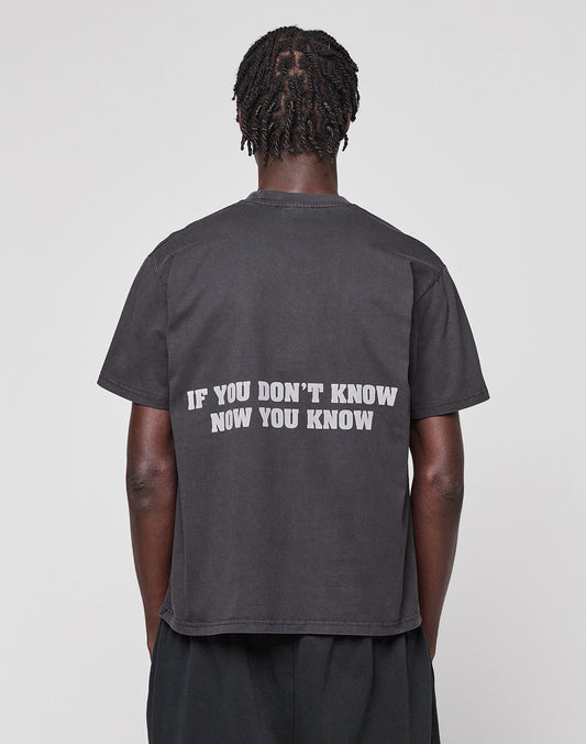 A person with short, twisted hair is seen from the back wearing the LIVE FAST DIE YOUNG Abus Magomedov Tee—a black regular fit t-shirt with "IF YOU DONT KNOW NOW YOU KNOW" in white as a backprint. The background is plain and light-colored.