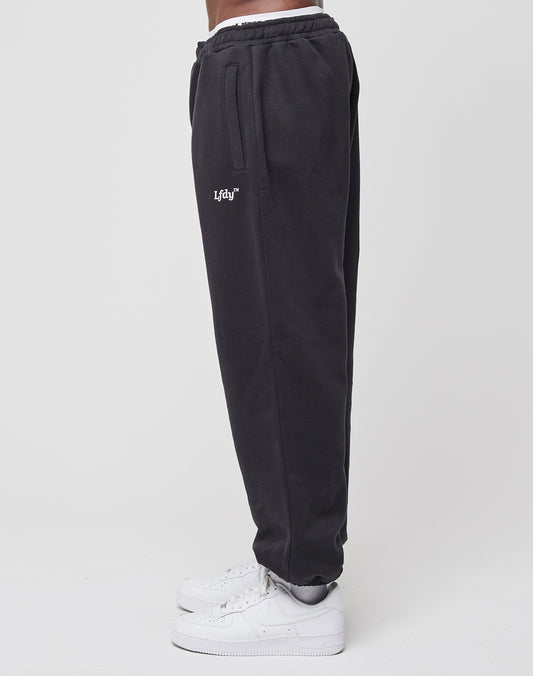 A person stands sideways against a plain white background, wearing black LIVE FAST DIE YOUNG Loose Jogger sweatpants with an elastic waistband and Lfty logo, paired with white sneakers.