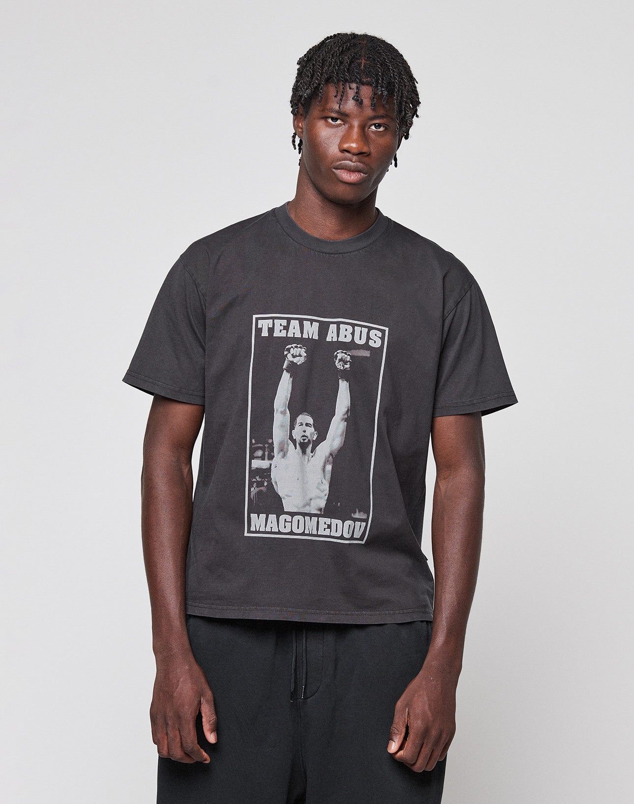 A young man with short dreadlocks wears the Abus Magomedov Tee by LIVE FAST DIE YOUNG—100% Baumwolle, black, featuring a victorious athlete graphic and "TEAM ABUS MAGOMEDOV" text—against a plain light gray background.