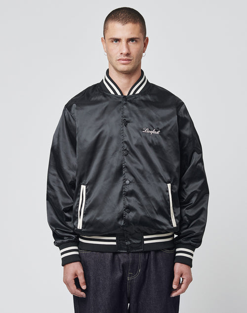 A man faces forward, wearing the LIVE FAST DIE YOUNG Satin College Jacket with white striped trim and Logo-Stick embroidery on the left chest, paired with dark jeans, against a plain light gray background.