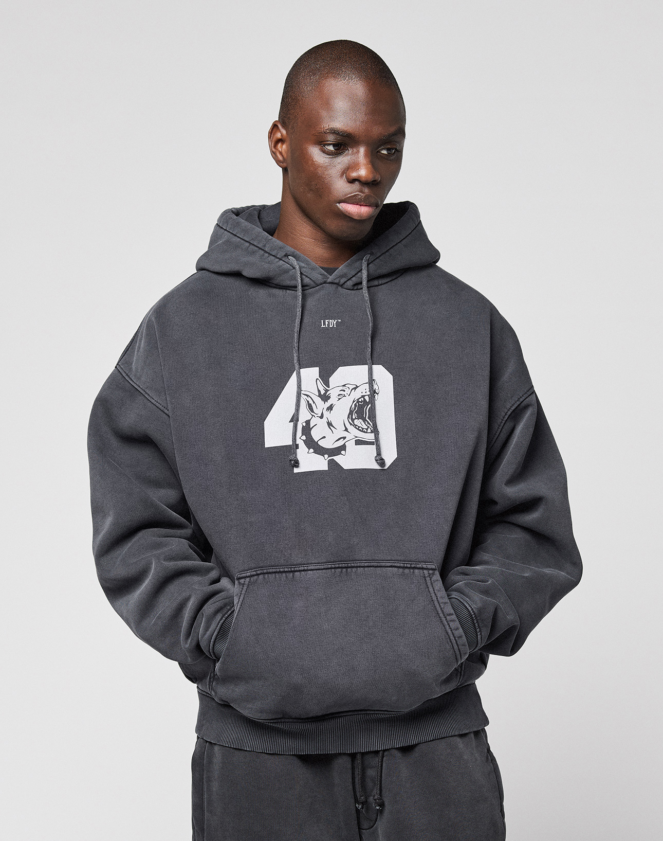 A young man wears the LFDY 40 Hoodie by LIVE FAST DIE YOUNG, an oversized hoodie with drawstrings, a white graphic, and a Kangaroo-Tasche, standing against a plain light gray background with hands in the front pocket.