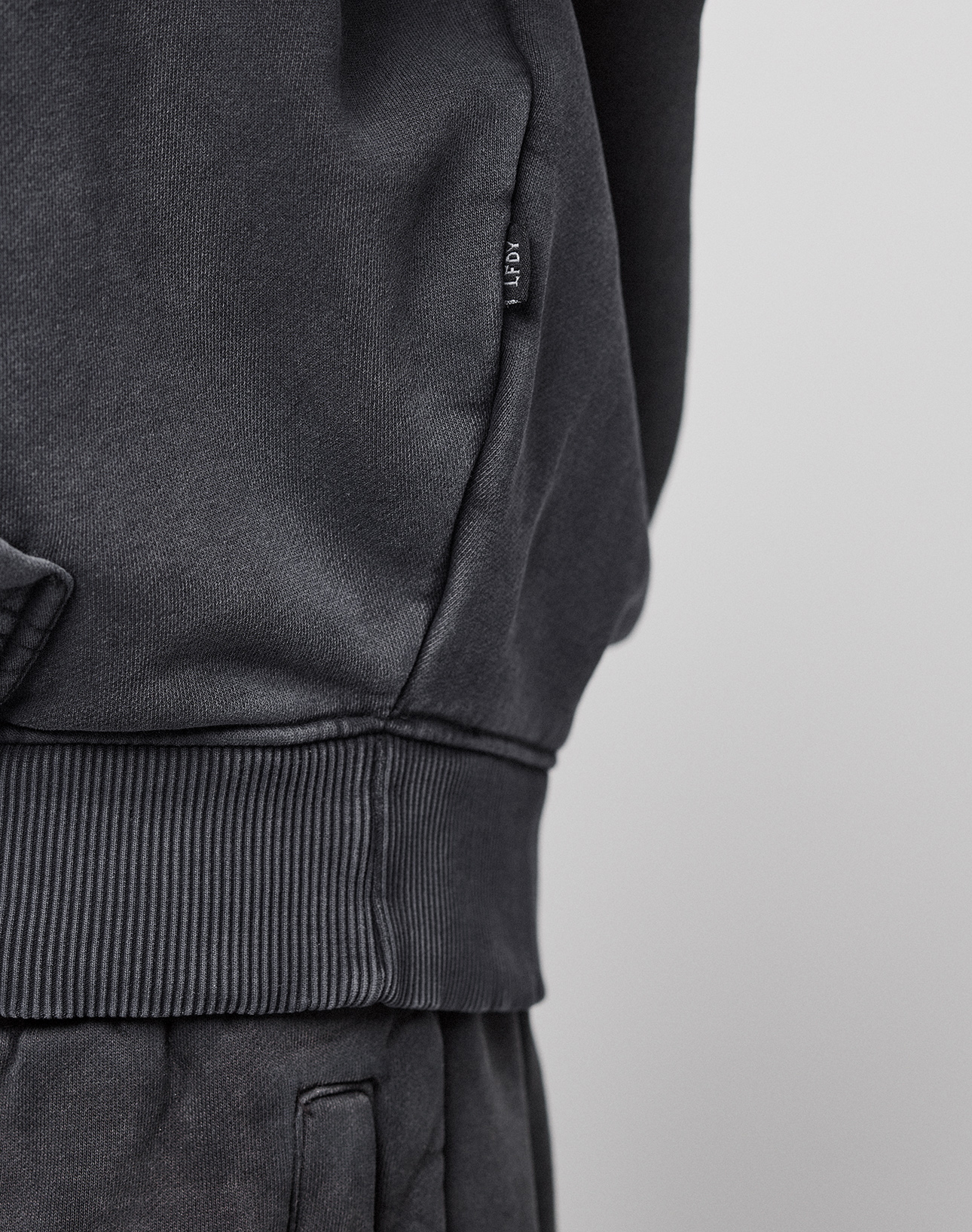 A close-up shows a person wearing the dark grey LFDY 40 Hoodie by LIVE FAST DIE YOUNG with matching pants, highlighting the ribbed waistband and a small tag on the side seam. The neutral background is out of focus.