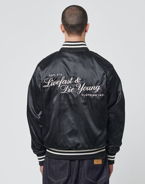 A person with short hair, seen from behind, wears the LIVE FAST DIE YOUNG Satin College Jacket featuring white striped cuffs/collar, Druckknöpfe, a subtle logo stick, and “Live Fast & Die Young” script on the back. | model