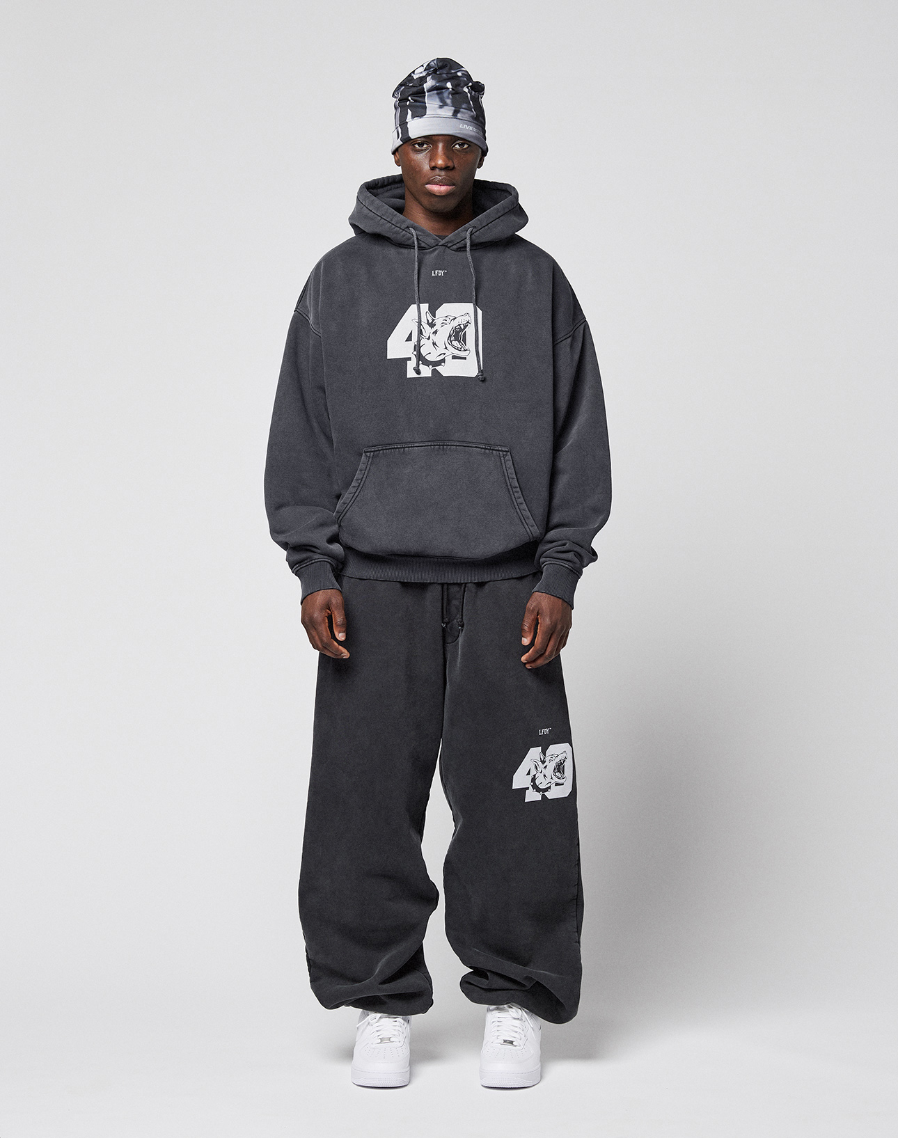 A person stands facing forward in a dark LFDY 40 Hoodie and matching sweatpants by LIVE FAST DIE YOUNG, both featuring "40" graphics, paired with white sneakers and a patterned headscarf against a plain light gray background.