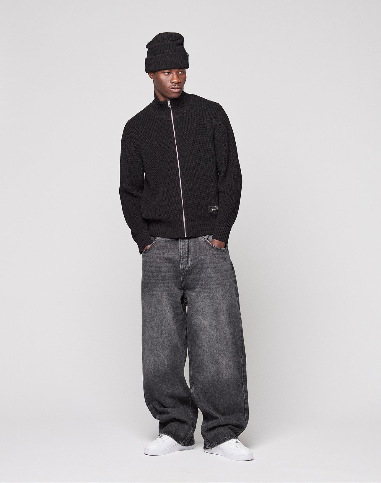 A person in a black LFDY Knit Jacket by LIVE FAST DIE YOUNG, oversized faded black 100% cotton jeans, black beanie, and white sneakers stands with hands in pockets against a plain light background.