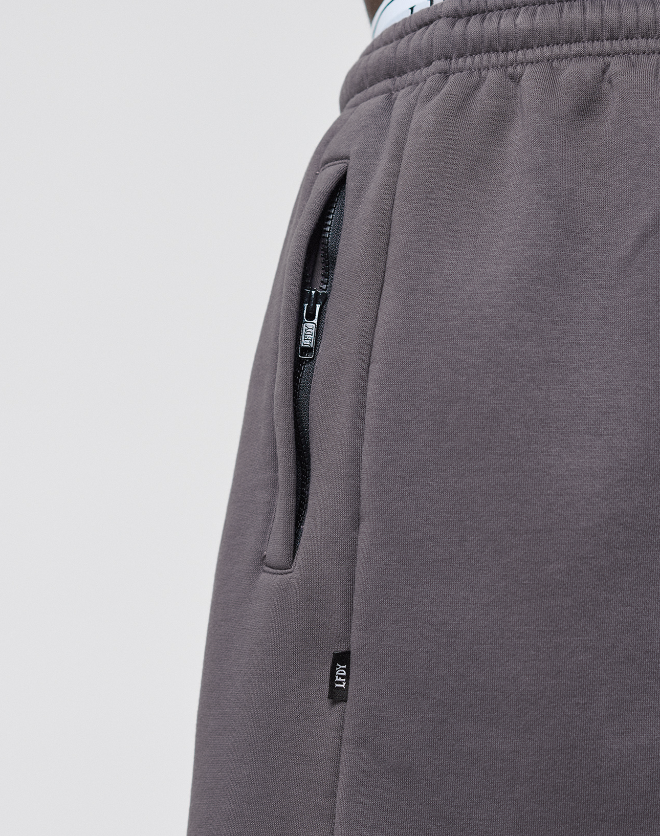 Close-up of a person wearing LIVE FAST DIE YOUNG's Live Fast LF Shorts in dark gray, featuring elastic waistband, wide fit, side slit pockets with a zippered pocket, and a small black tag with white text near the seam.