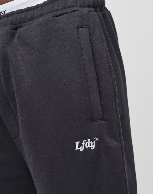 Close-up of black LIVE FAST DIE YOUNG Loose Jogger with white LfdY™ embroidery near the pocket, seitliche Eingrifftaschen, and Elastik-Bund. The waistband of white underwear peeks slightly above the pants.