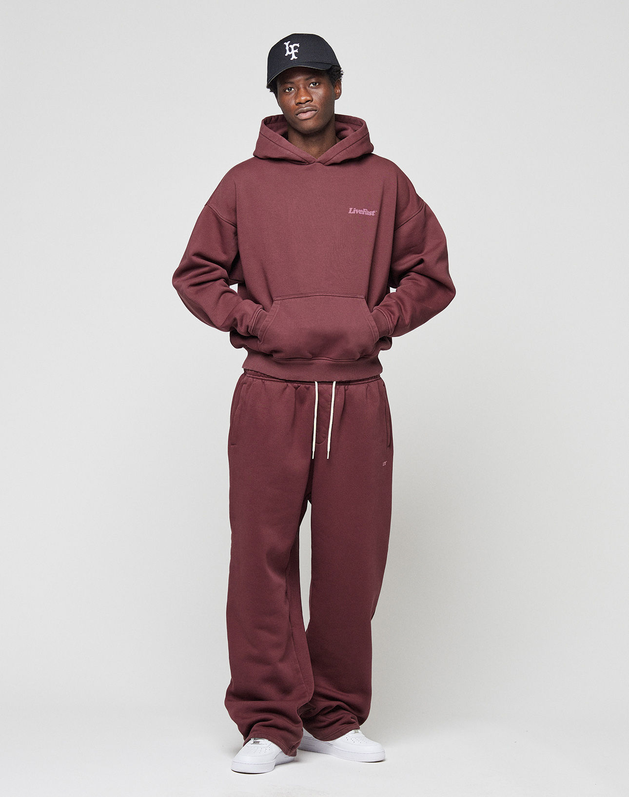 A person stands against a plain background wearing the LIVE FAST DIE YOUNG Basic Wide Jogger hoodie and sweatpants in burgundy, paired with a black cap and white sneakers, with hands tucked into the hoodie’s front pocket.