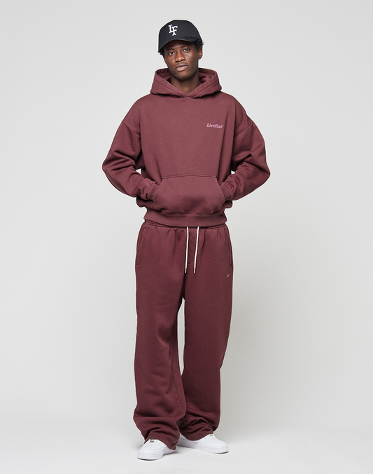 A person stands against a plain background wearing the LIVE FAST DIE YOUNG Basic Wide Jogger hoodie and sweatpants in burgundy, paired with a black cap and white sneakers, with hands tucked into the hoodie’s front pocket.