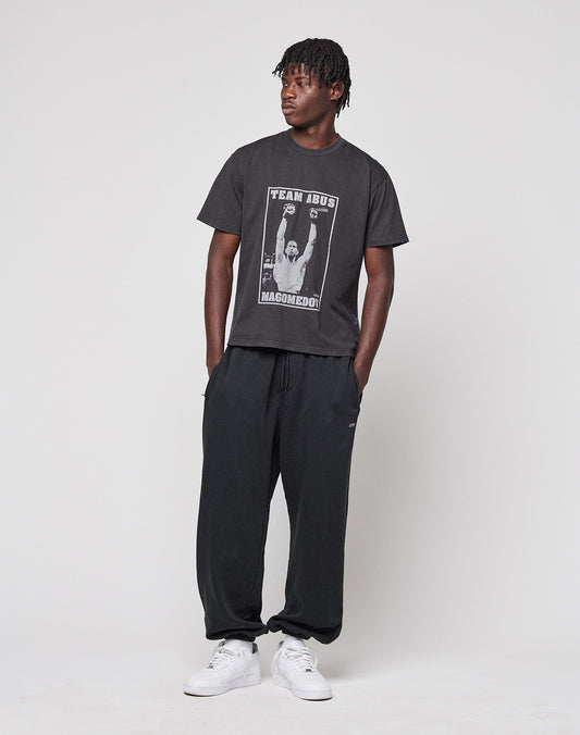 A person with short, twisted hair stands against a plain background, wearing the Abus Magomedov Tee by LIVE FAST DIE YOUNG—a Regular Fit black T-shirt made of 100% Baumwolle with photo and text prints on front and back—plus black sweatpants and white sneakers.