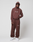 A person stands against a plain background wearing a matching brown French Terry hoodie and LIVE FAST DIE YOUNG Wide Cuff Jogger with white lettering and sneakers. One hand is in their pocket as they look at the camera.
