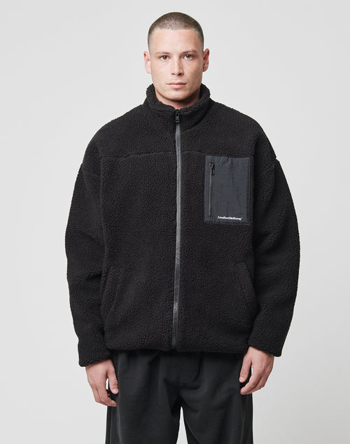 A person with short hair wears the LIVE FAST DIE YOUNG Basic Teddy Jacket, a regular fit black zip-up fleece with Stehkragen and Brusttasche, standing against a plain light gray background.