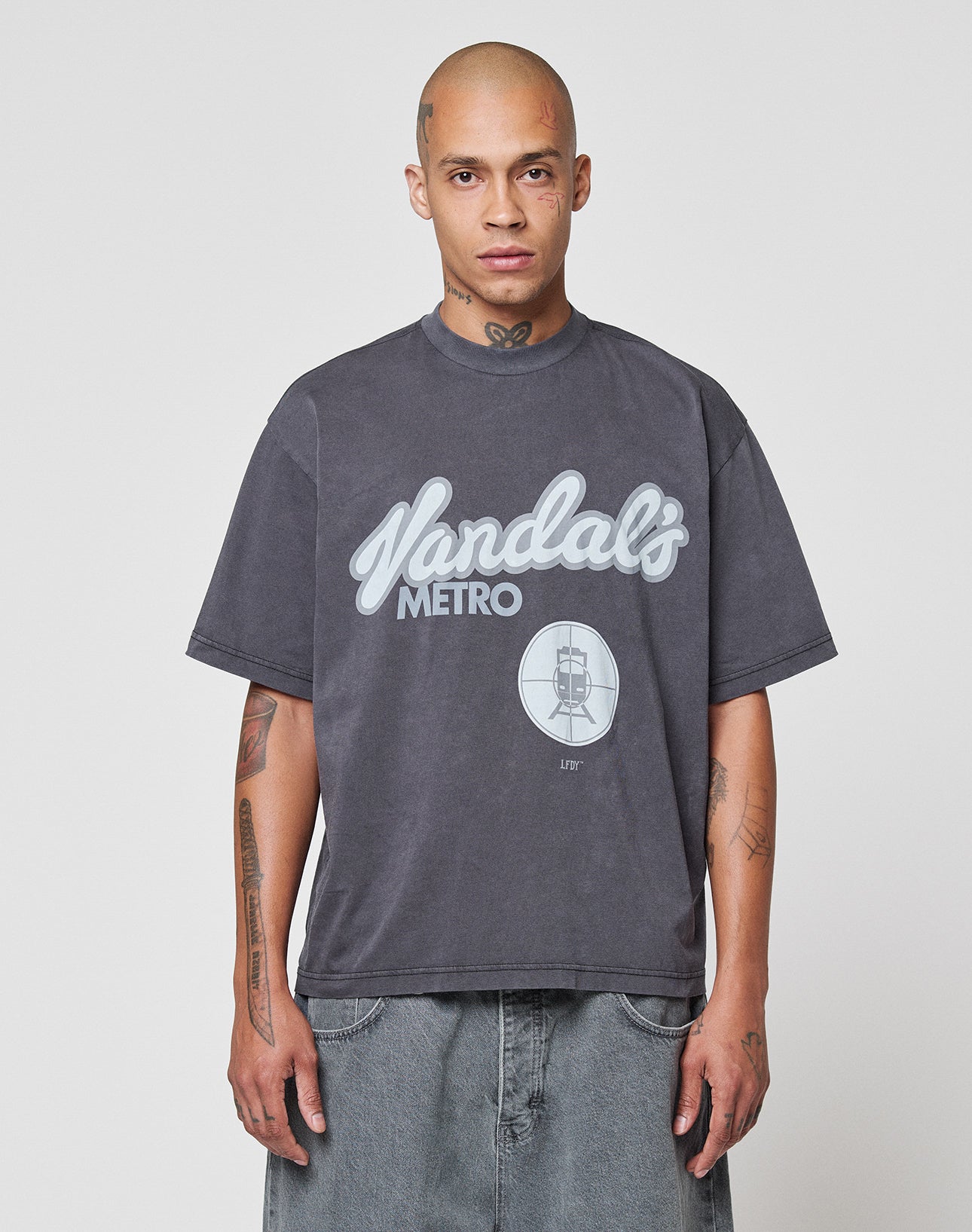 A person with a shaved head and tattoos wears the LIVE FAST DIE YOUNG LFDY Vandals Tee—an oversize fit, 100% cotton dark gray shirt with a Metro frontprint and train graphic—paired with baggy denim shorts against a white background.