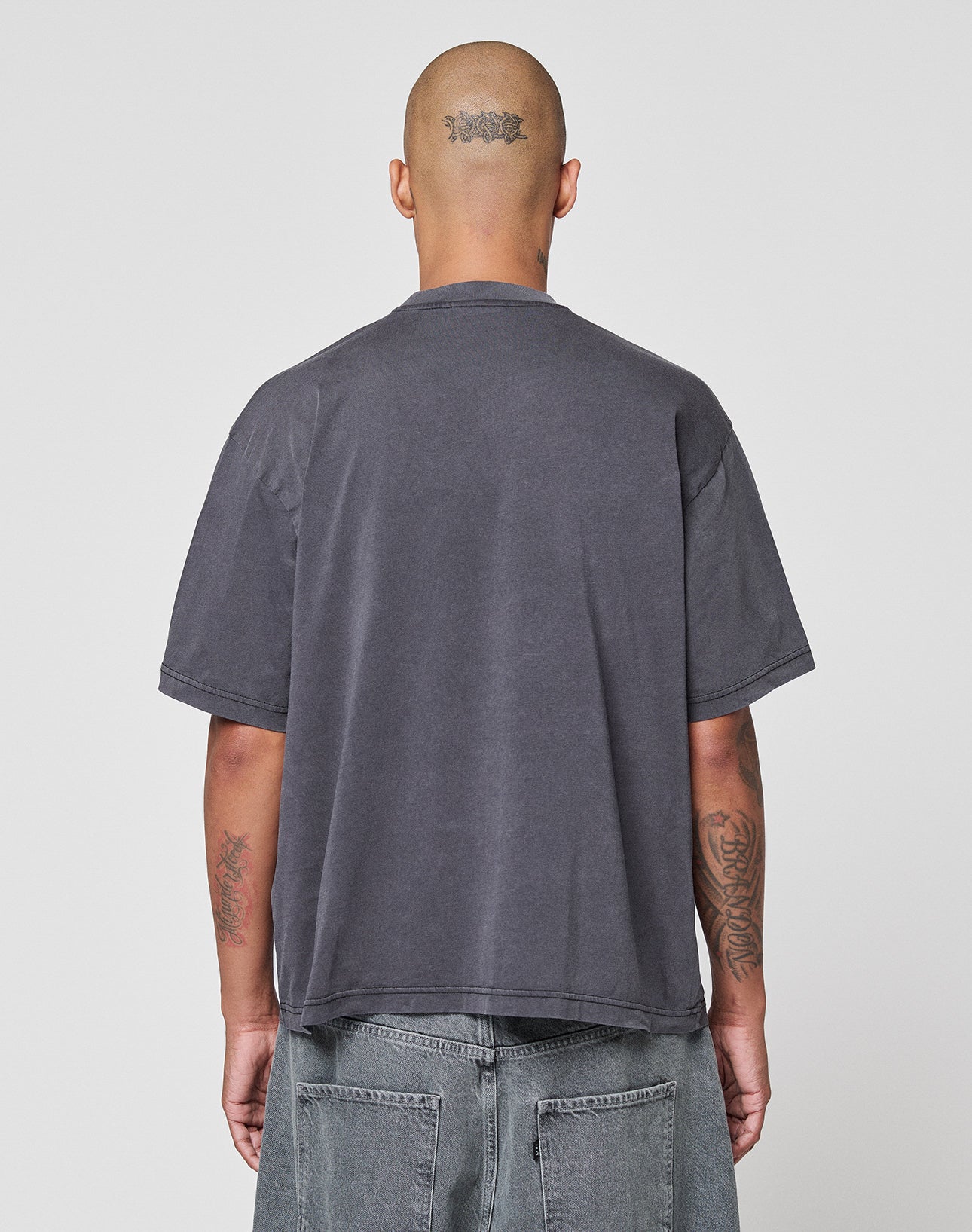 A person with a shaved head and visible tattoos wears an oversize fit, faded dark gray LFDY Vandals Tee by LIVE FAST DIE YOUNG and gray jeans, standing with their back to the camera against a plain white background.