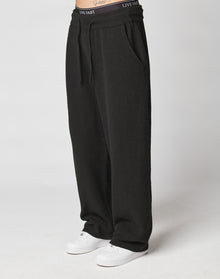 A person wears LIVE FAST DIE YOUNG Live Fast Boucle Pants—black, wide-fit, 100% polyester sweatpants with an elastic waistband printed LIVE FAST and side pockets, styled with white sneakers against a plain light background.