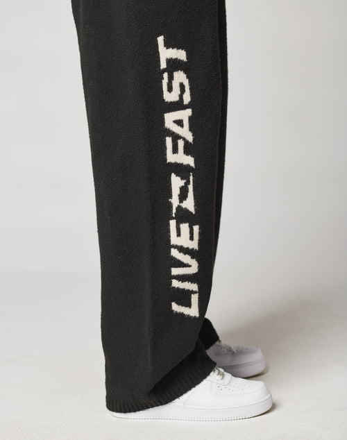 A person wears the Live Fast Boucle Pants by LIVE FAST DIE YOUNG—wide fit, black, with bold white LIVE FAST lettering down the side and an elastic waistband—styled with white sneakers against a light background.