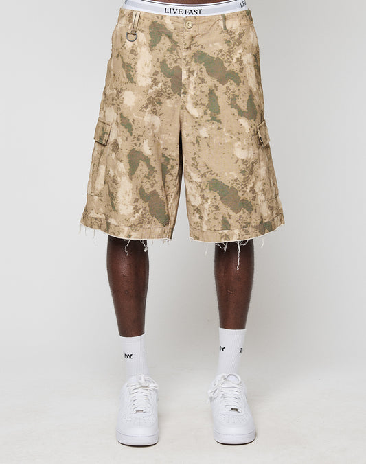 A person wears LFDY Cargo Shorts by LIVE FAST DIE YOUNG in tan and green camo with frayed hems, paired with a white LIVE FAST waistband, white socks, and white sneakers, showing a vintage style against a plain white background.