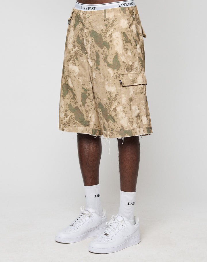 A person wears beige and green LFDY Cargo Shorts by LIVE FAST DIE YOUNG, white LIVE FAST waistband underwear, white LFIV socks, and white sneakers. Only the lower body is visible against a plain background. | model