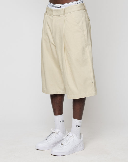 A person wears beige LFDY Twill Shorts by LIVE FAST DIE YOUNG made from 100% Baumwolle with weites gerades Bein, plus white socks and underwear reading LIVE FAST, and white Nike sneakers, all against a plain white background.