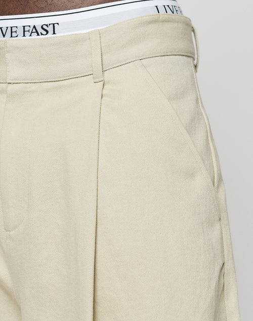 Close-up of a person wearing beige LFDY Twill Shorts by LIVE FAST DIE YOUNG, featuring a wide straight leg and belt loops. The waistband of white underwear with “LIVE FAST” in black is visible above the 100% cotton shorts.
