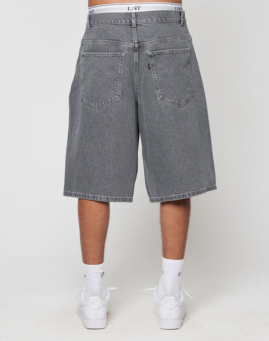 A person faces away wearing LIVE FAST DIE YOUNG LFDY Jorts, knee-length gray denim shorts in Baggy Fit with weites gerades Bein, paired with white socks, sneakers, and a visible white waistband. The background is light and plain.