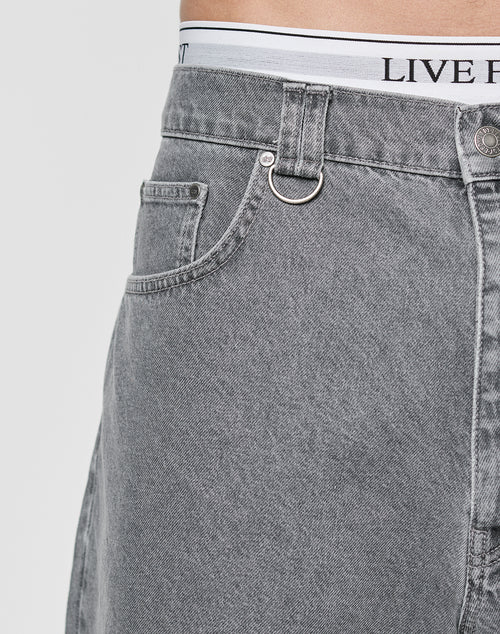 Close-up of a person’s waist wearing gray LFDY Jorts by LIVE FAST DIE YOUNG. These 100% Baumwolle baggy-fit jeans feature a front pocket, small loop, visible metal button, and a white waistband reading LIVE FAST.
