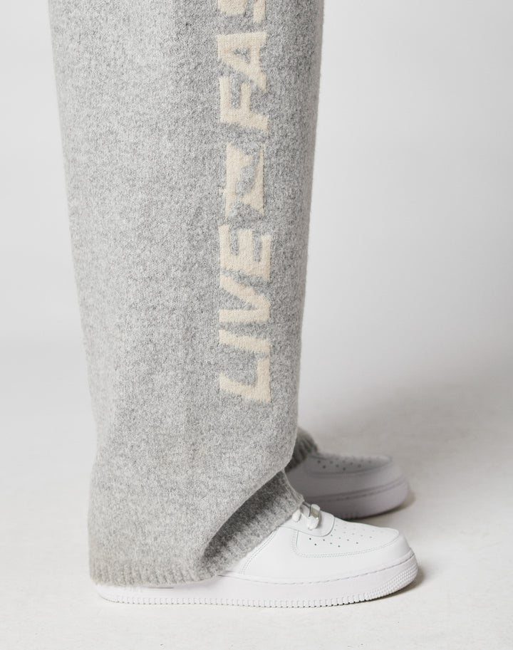 A person wearing LIVE FAST DIE YOUNG’s Live Fast Boucle Pants—wide fit gray sweatpants with beige “LIVE FAST” text and an elastic waistband—paired with white sneakers, stands on a light-colored surface.