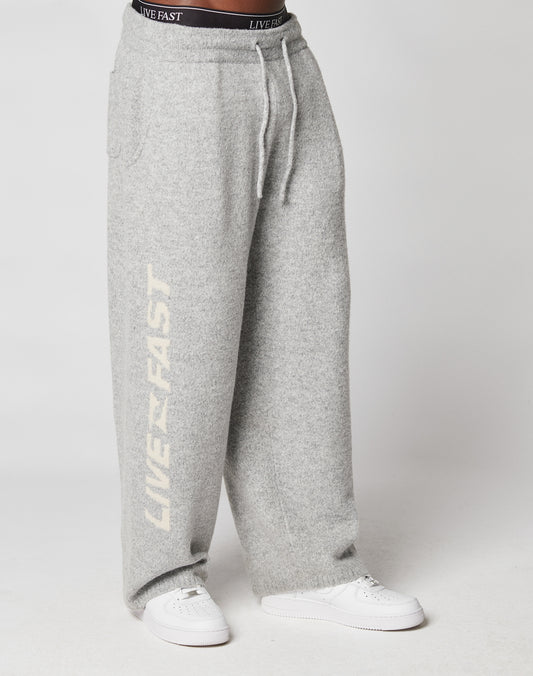Someone wears loose-fitting, light gray Live Fast Boucle Pants by LIVE FAST DIE YOUNG, featuring an elastic waistband and LIVE FAST in white on one leg, styled with white sneakers and a visible black LIVE FAST waistband.