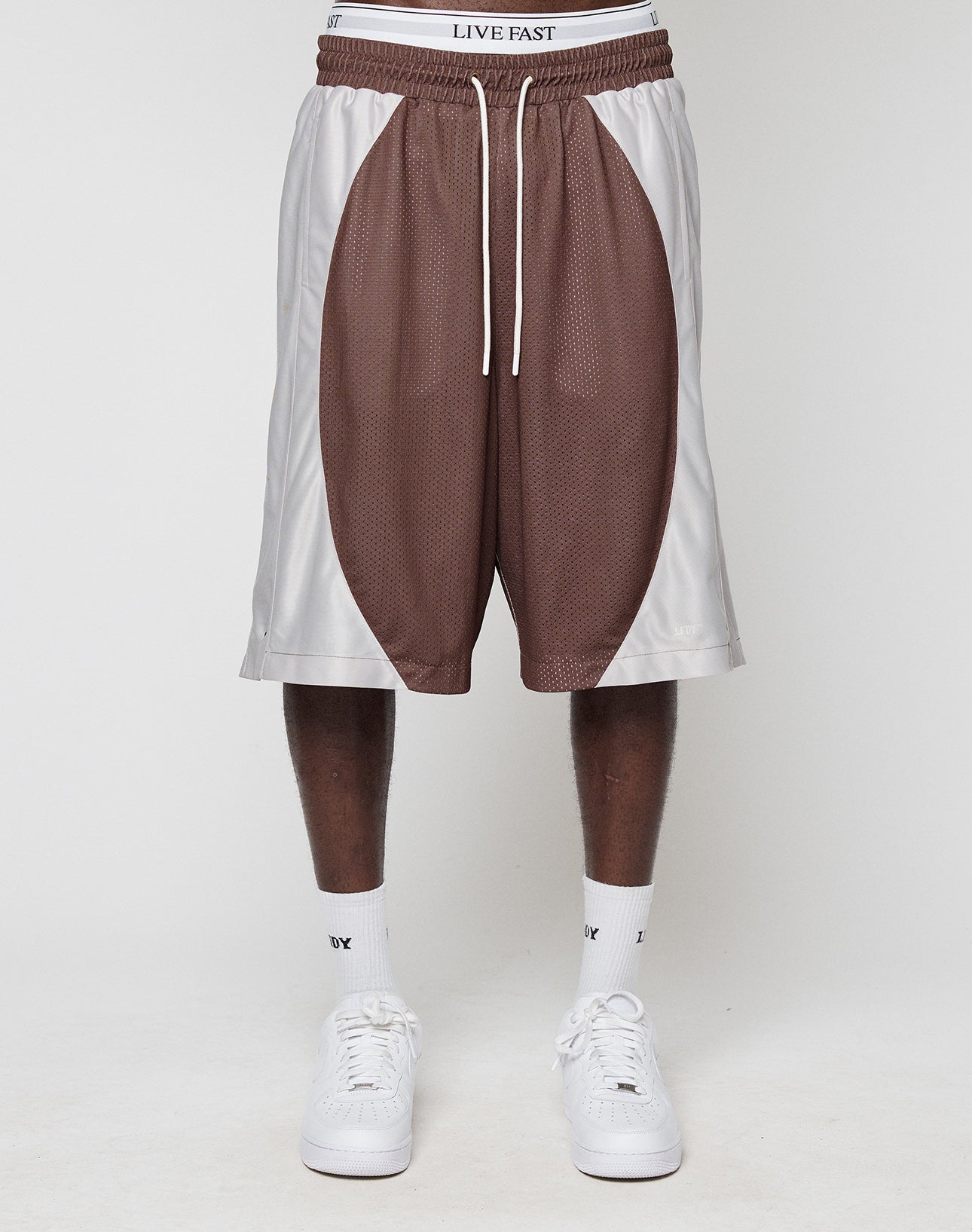 A person is shown from the waist down wearing LIVE FAST DIE YOUNG LFDY League Shorts in brown and white, paired with white socks and sneakers, standing against a plain white background.