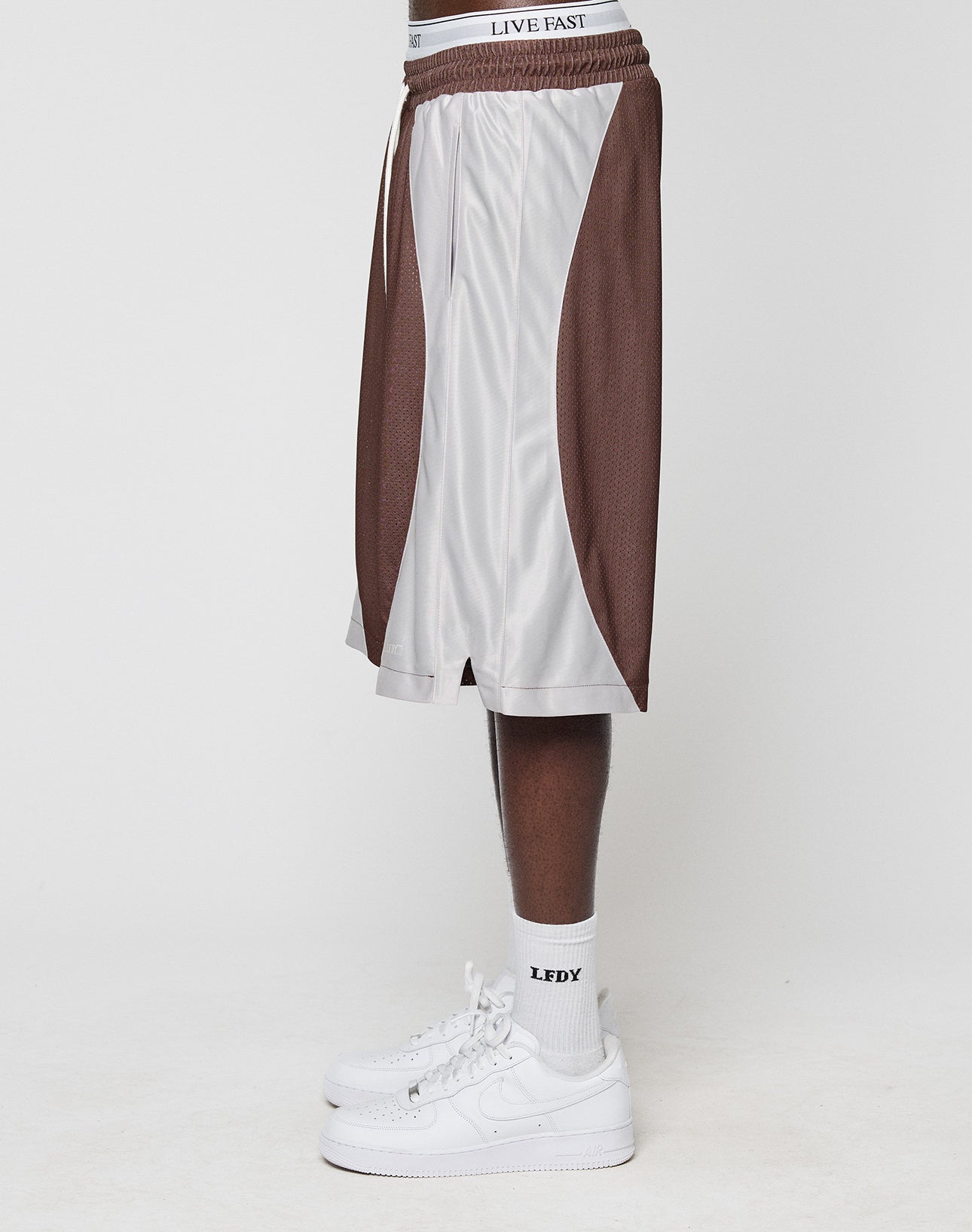 A person stands sideways in brown and white LFDY League Shorts by LIVE FAST DIE YOUNG, with white LEDY socks and white Nike sneakers. Only their lower half is visible against a plain background.