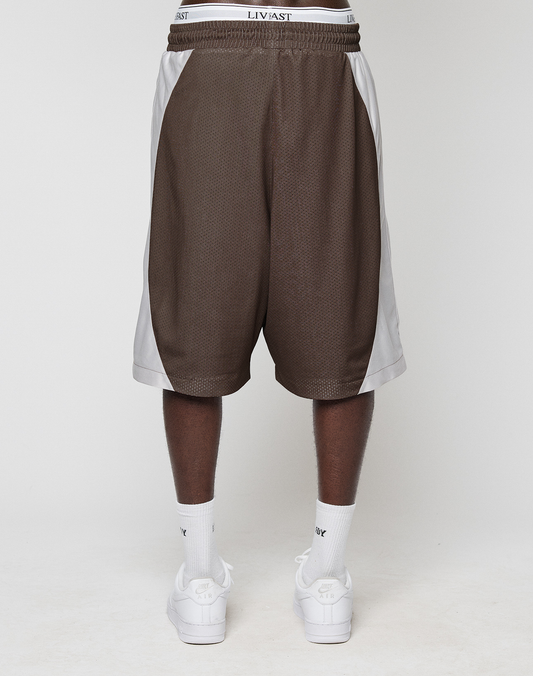 A person stands facing away from the camera wearing oversize brown and white LIVE FAST DIE YOUNG LFDY League Shorts with a bold logo-print waistband, paired with white Nike socks and white sneakers.