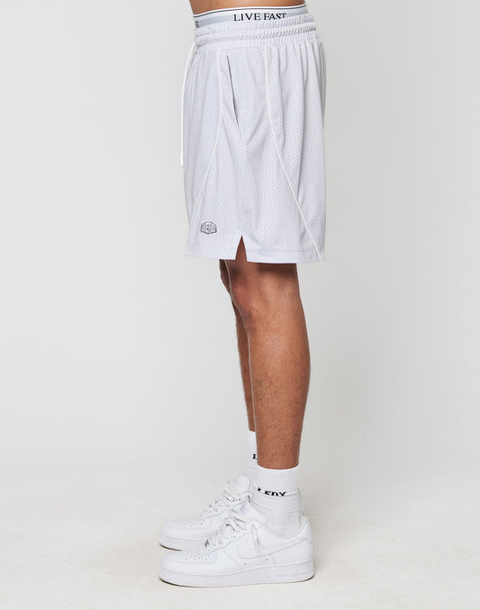 A person stands sideways in LFDY Piping Mesh Shorts by LIVE FAST DIE YOUNG—regular fit, white polyester with small side slit and elastic drawstring waist—paired with white crew socks and sneakers, against a plain white background.
