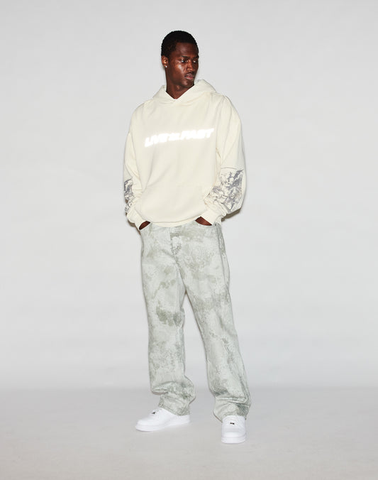 A man wears the LFDY Window Hooded by LIVE FAST DIE YOUNG, featuring “LATE FAST” reflective print, logo embroidery, patterned sleeves, light grey patterned pants, and white sneakers. He looks down left for an oversized fit style.
