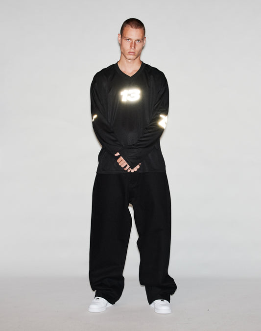 A person stands against a plain light background, wearing the LIVE FAST DIE YOUNG Live Fast Jersey—an oversized black long-sleeve shirt with a reflective 13 print on the chest—wide black pants, and white sneakers, looking at the camera with hands clasped.