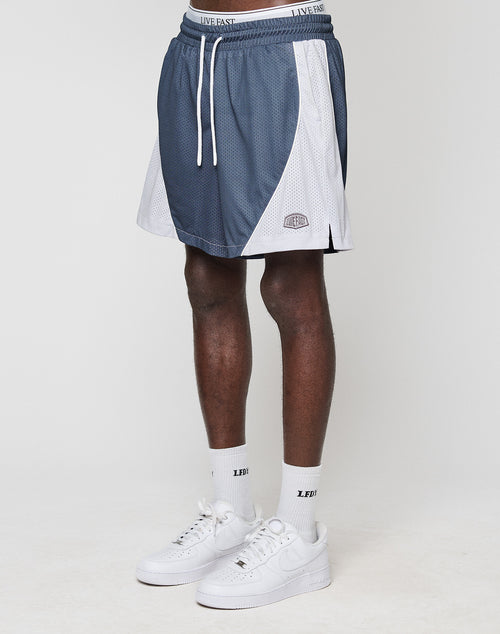 A person wears blue and white LFDY Piping Mesh Shorts by LIVE FAST DIE YOUNG with a LIVE FAST waistband, white socks with LED text, and white Nike sneakers, standing against a plain white background.