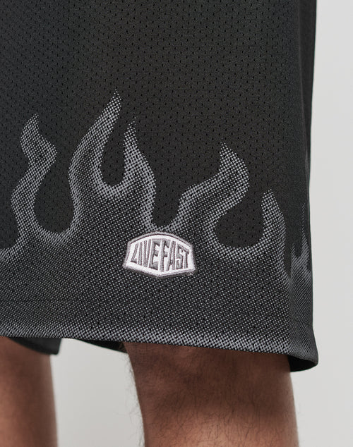 Close-up of the Burning Mesh Shorts by LIVE FAST DIE YOUNG, featuring black mesh fabric, a gray flame print, and a LIVE FAST patch. The oversize fit with elastic waistband ensures comfort; part of a leg is visible at the bottom.