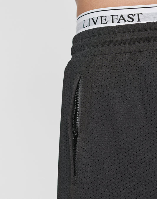 Close-up of a person wearing Burning Mesh Shorts by LIVE FAST DIE YOUNG, featuring an elastic waistband and zippered pocket. The white underwear waistband with LIVE FAST is visible above the shorts.
