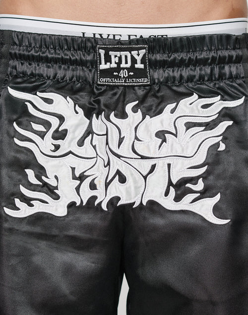 Close-up of LFDY Fighting Shorts from LIVE FAST DIE YOUNG: black satin, elastic waist with LIVE FAST in white, bold flame-like LIVE FAST embroidery on the back, and a black patch reading LFDY 40 OFFICIALLY LICENSED. Regular fit.