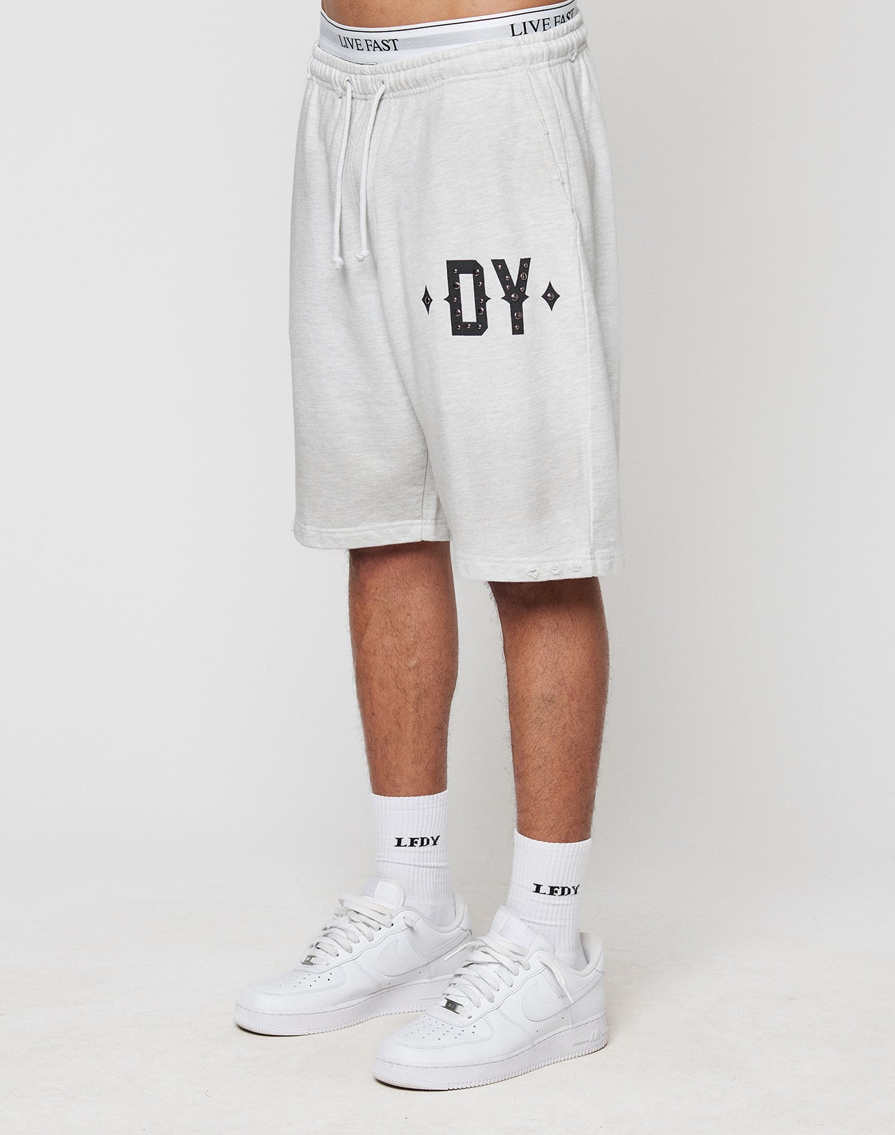 A person in LIVE FAST DIE YOUNG DY Rivet Shorts (light gray, 100% Baumwolle) with DY on the left leg, white LFDY socks, and white Nike sneakers stands against a plain white background.