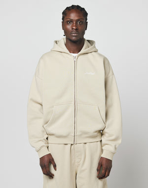 A person with braided hair wears a beige LIVE FAST DIE YOUNG Basic Script Hooded Zip and matching pants, standing against a plain light gray background.