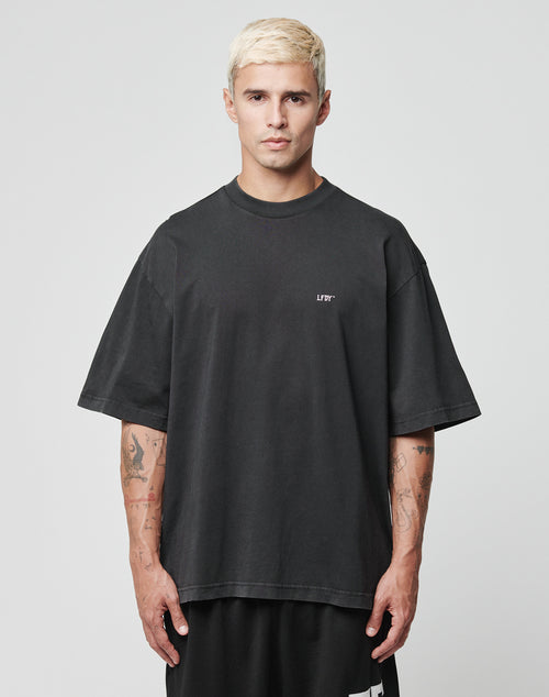A man with short bleached blonde hair wears the LIVE FAST DIE YOUNG Everybody Eats Tee, an oversize black 100% Baumwolle T-shirt, featuring logo on the chest. He stands facing forward against a plain light background, arms relaxed to show tattoos.