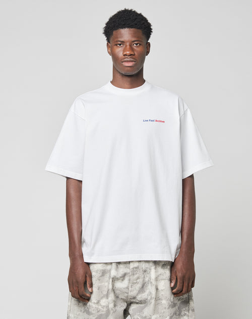 A man stands facing forward wearing the LIVE FAST DIE YOUNG LFDY F*ck Phones Tee, a 100% Baumwolle plain white Oversize Fit T-shirt with small red text on the upper chest, and light gray camo-patterned pants against a light gray background.